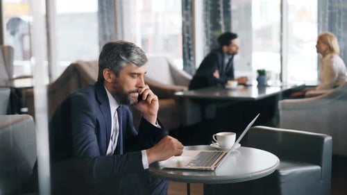 Serious Businessman Speaking on Mobile Phone and Working with Laptop in Cafe