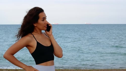 Beautiful tanned girl in sportswear talking on the phone with a sea view.