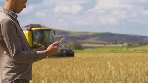Farmer Using Tablet While Tractor Harvests Crops