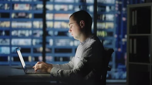 Nachtarbeit: Ein Mann sitzt an einem Tisch und druckt auf einem Laptop vor dem Hintergrund der Nachtstadt