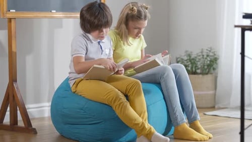 Wide Shot of Caucasian Boy and Girl Sitting on Comfortable Bag Chair in Classroom Reading Books