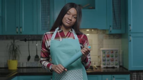 Young African American Woman in Apron with Kitchen Utensil in Hands Standing on Blue Kitchen Looking
