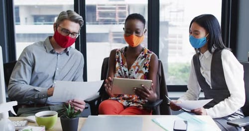 Diverse office colleagues wearing face masks discussing together at modern office