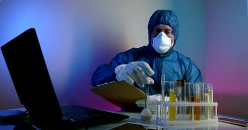 Un hombre con ropa protectora con capucha, guantes y un respirador está sentado en una mesa con una prueba