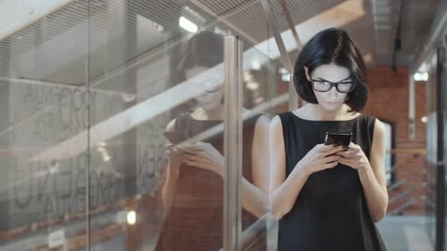 Stylish Businesswoman Leaning on Glass Office Wall and Texting on Phone