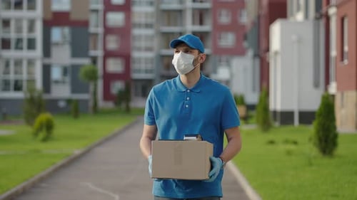 The Male Deliveryman in a Cap and a Protective Mask and Gloves Goes with a Box in His Hands and