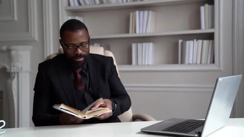 Black Man Is Working in His Home Office, Reading Book, Sitting at Table with Laptop