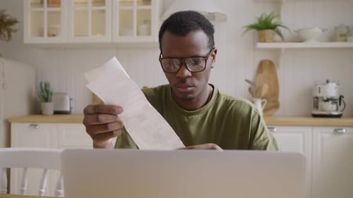 African American Man Looks at Checks Near Laptop in Kitchen