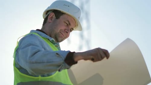 Man in Hardhat Inspecting Plans Near Power Lines