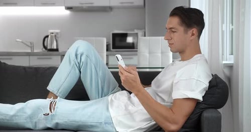 Close Up of Cheerful Young Man Resting on the Sofa Using Smartphone Smile in the Modern Apartment