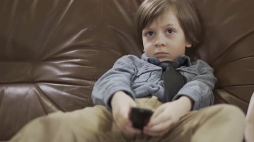 Der kleine süße Junge sitzt mit gespreizten Beinen auf dem Ledersofa und wechselt mit der Fernbedienung die Kanäle im Fernsehen