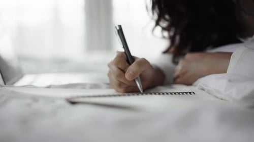 close-up woman writing notebook and using laptop computer on a bed