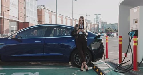 Frau steht mit Telefon in der Nähe ihres Elektroautos und wartet, wann das Fahrzeug wird