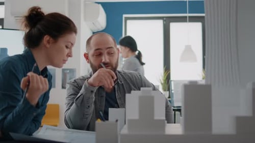 Man and Woman Pointing at Building Model to Plan Blueprints on Paper