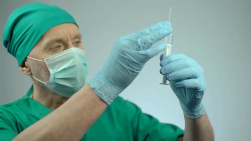 Anesthesiologist in Face Mask Checking Syringe for Injection, Operating Theater