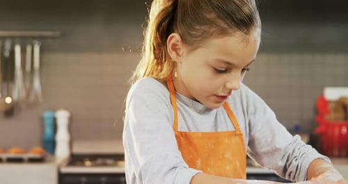 Concentrated little girl wearing apron and kneading dough at kitchen
