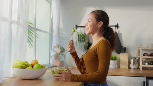 Attractive young Latino woman eating green salad in kitchen at home.