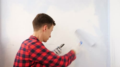 Young Adult Painting Wall While Using Smartphone