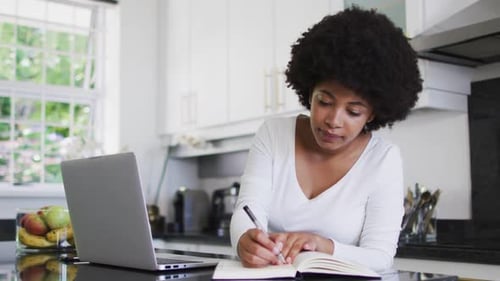 Woman Writes in Notebook with Laptop in Kitchen