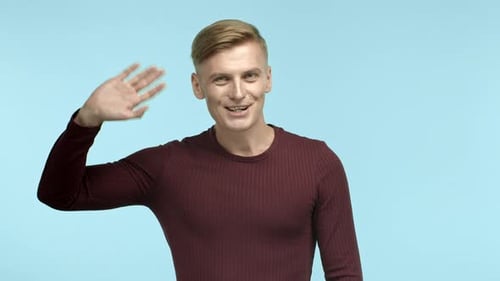 Handsome Blonde Man Saying Hello and Waving Hand at Camera Greeting you and Smiling Standing Over