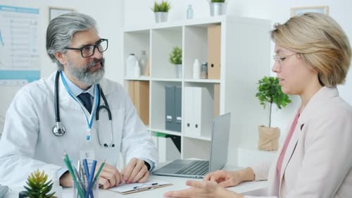 Friendly Mature Doctor Talking to Female Patient During Appointment in Clinic Discussing Disease and