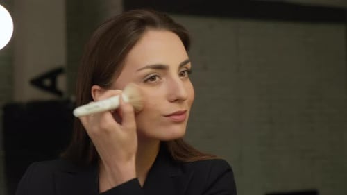 beautiful young woman in bathrobe using cosmetic brush in front of mirror