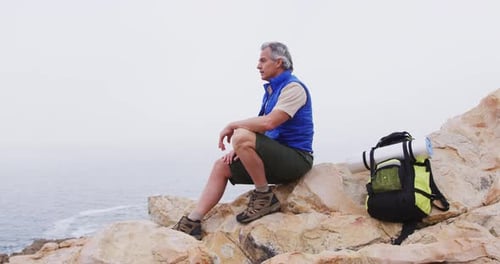 Senior hiker man with backpack sitting on the rocks in the mountains and watching the sea shore.