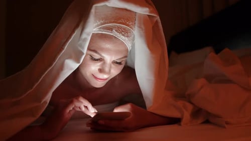 Beautiful Young Woman Using Smart Phone Under Blanket in Bedroom on Bedtime