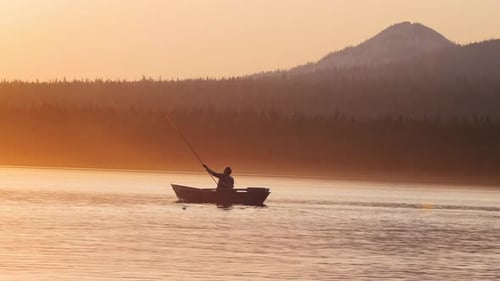 Eine Silhouette eines Mannes, der in einem Boot sitzt - Angeln in der Natur während des Sonnenuntergangs