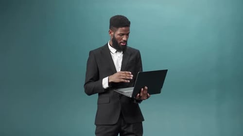 Ein attraktiver afrikanischer bärtiger Mann steht in einem Smoking, hält einen Laptop in den Händen und spricht