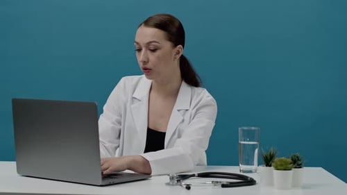 Sexy Female Doctor in a White Medical Coat Sits at a Table and Works at Laptop