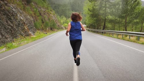 Woman running on mountain road surrounded by green tree forest.