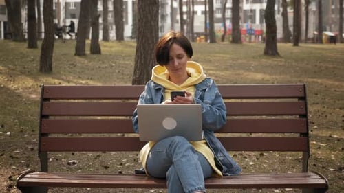 Young woman sitting on bench in spring park using laptop and smartphone
