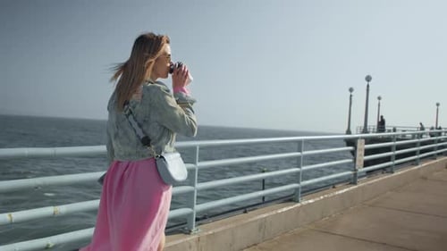 Stylish Happy Woman in Flowing Pink Dress Walking By Ocean Pier Drinking Coffee
