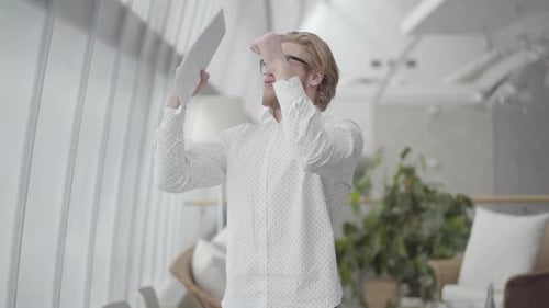 Blond Happy Man in Glasses Dancing in a Light Comfortable Office Holding Papers in Hands