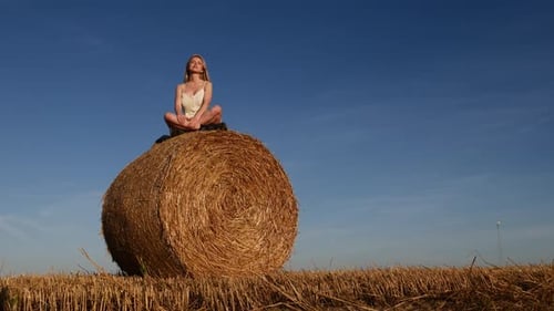 blonde girl is sitting on rolled haystack in field in sunset time