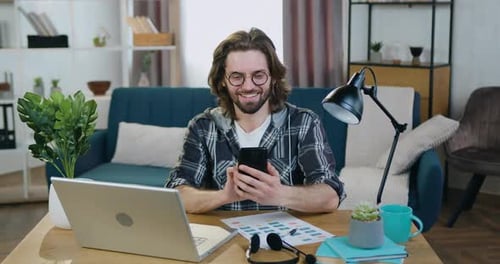 Mann mit Brille sitzt am Schreibtisch im gemütlichen Home Office und benutzt sein Smartphone