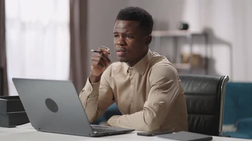 Thoughtful Black Man is Sitting at Table with Open Laptop and Trying to Concentrate Working Remotely