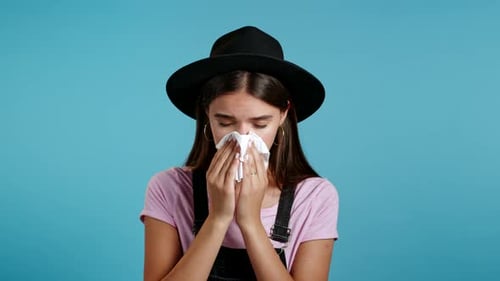 Hipster Girl Sneezes Into Tissue. Isolated Woman on Blue Studio Background Is Sick, Has a Cold or