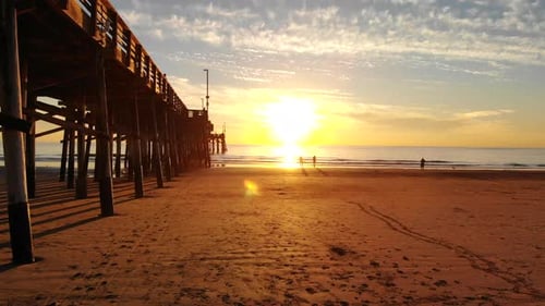 Drone flying under Newport Beach pier at sunset as sunlight beams and people walk the California coa