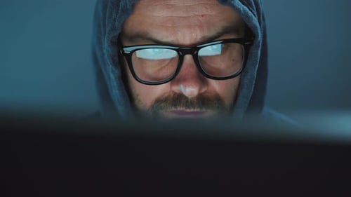 Male Hacker in the Hood and Glasses Working on a Computer in a Dark Office Room. Cybercrime Concept
