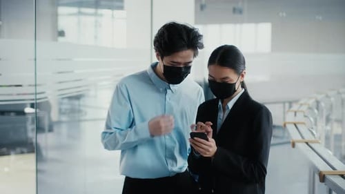 Two Japanese Coworkers Using Smartphones Talking Wearing Masks In Office