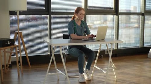 Cheerful Young Female Doctor Wearing Blue Green Medical Uniform Typing on Laptop Computer Looking on
