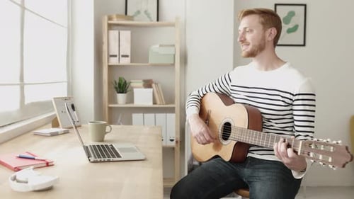 Young Man Playing Guitar at Home by Desk