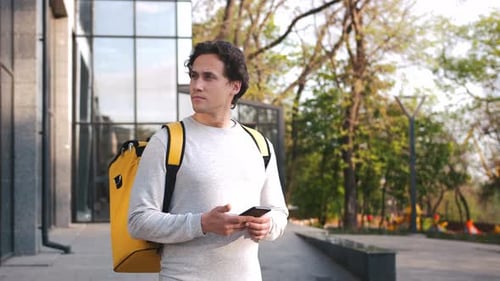 Porträt eines kaukasischen Zustellers, der mit gelbem Rucksack steht, mithilfe der Navigations-App am Telefon