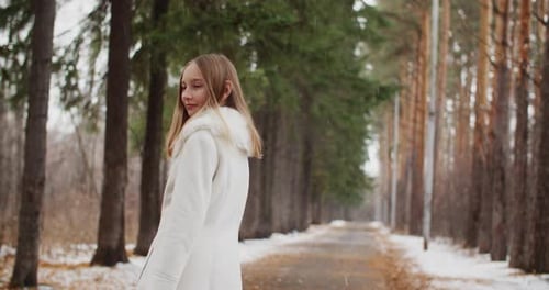 Attractive Young Girl in White Coat with Fur Collar Looking Back and Shaking Hair on Tree Alley at
