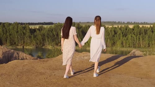 Two Slender Women in White Dresses Walk to Edge of Abyss in Mountains with Beautiful View of Blue