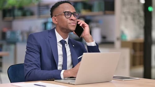 African Businessman with Laptop Talking on Smartphone in Office