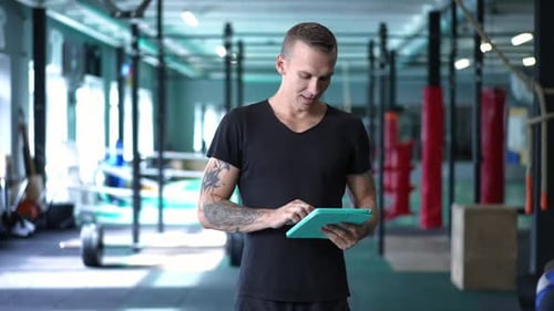 Tattooed Handsome Caucasian Man Standing in Gym Surfing Internet on Tablet and Smiling Looking at