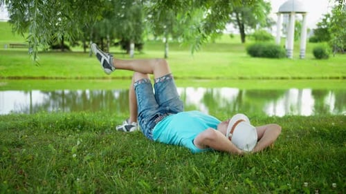 Happy young man in hat resting lying on the grass in city park under a tree